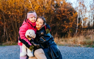 Mother in black leather coat kneels to hug her 7-year-old daughter with special needs, who holds white teddy bear. Golden sunset light and autumn trees create emotional and touching family portrait.