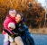 Mother in black leather coat kneels to hug her 7-year-old daughter with special needs, who holds white teddy bear. Golden sunset light and autumn trees create emotional and touching family portrait.